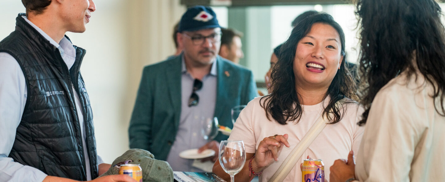 A casual networking event featuring three people engaging in a lively conversation around a tall table. The man on the left, wearing a black quilted vest, holds a can while smiling. The woman in the center, wearing a light shirt, smiles warmly as she speaks, with a can of LaCroix and an empty wine glass in front of her. Another person stands to the right, partially visible, listening attentively. The background includes other attendees, one wearing a teal blazer and cap, creating a relaxed and professional atmosphere.
