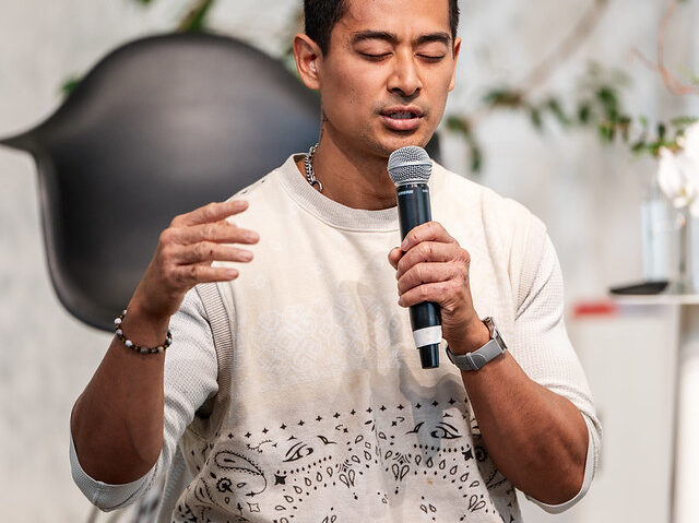 A man leading a guided meditation session, seated with his eyes closed and holding a microphone. He gestures gently with one hand, exuding calm and focus. Dressed in a patterned white sweater and camouflage pants, he creates a relaxed and grounded atmosphere. The background features a minimalist setup with greenery and a modern black chair, complementing the serene and mindful environment of the event.