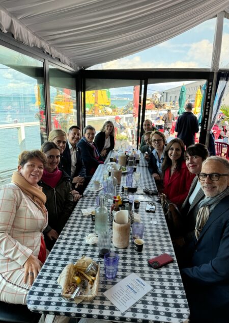 A group of people seated at a long table covered with a checkered tablecloth inside a covered patio area overlooking a harbor. The table is set with plates, glasses, and condiments, and colorful umbrellas and boats are visible through the windows in the background.