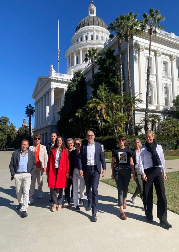 A group of individuals walking outside the California State Capitol building, characterized by its large dome and white exterior. The day is sunny, and the group is walking along a paved path surrounded by greenery.