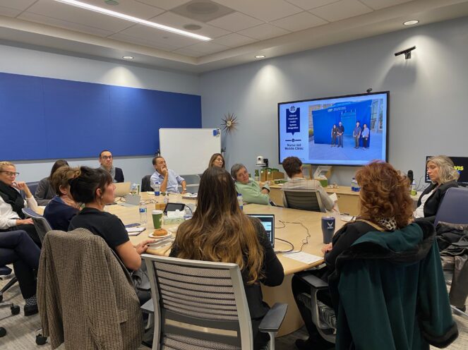 A group of people sitting around a conference table, watching a presentation on a large screen. The room features a modern design with blue accent walls and digital equipment.