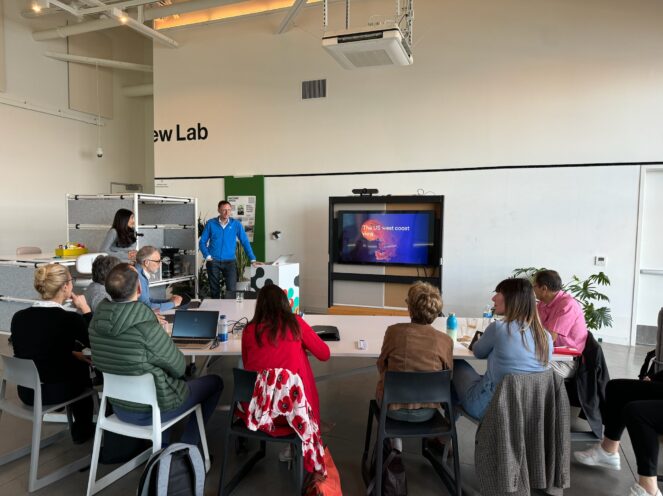 A group of individuals seated in a bright innovation lab, watching a presentation on a screen. The space has large windows, high ceilings, and modern office furniture.