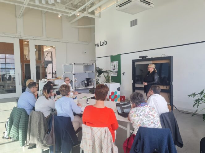 A group of attendees sitting at tables in a modern office space, listening to a presentation led by a blonde woman dressed in black. The space features an open design with high ceilings and contemporary furnishings.