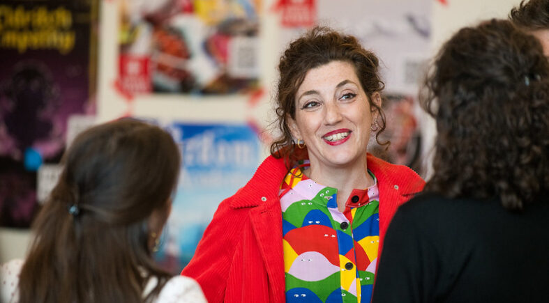 A vibrant scene featuring a woman in a colorful outfit with a cheerful expression, engaging in conversation with others in a lively environment. She wears a bright red jacket over a multicolored patterned shirt, exuding warmth and energy. The background displays posters and flyers, hinting at an event or community gathering focused on creativity or inclusivity. The atmosphere is lively and welcoming, fostering interaction and connection.