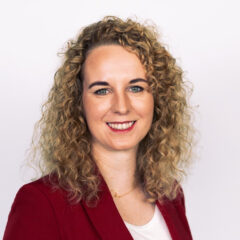 This image depicts a professional woman with curly blonde hair, wearing a red blazer over a white top, against a plain white background. She has a warm and confident smile, and her attire suggests a polished, business-appropriate look.