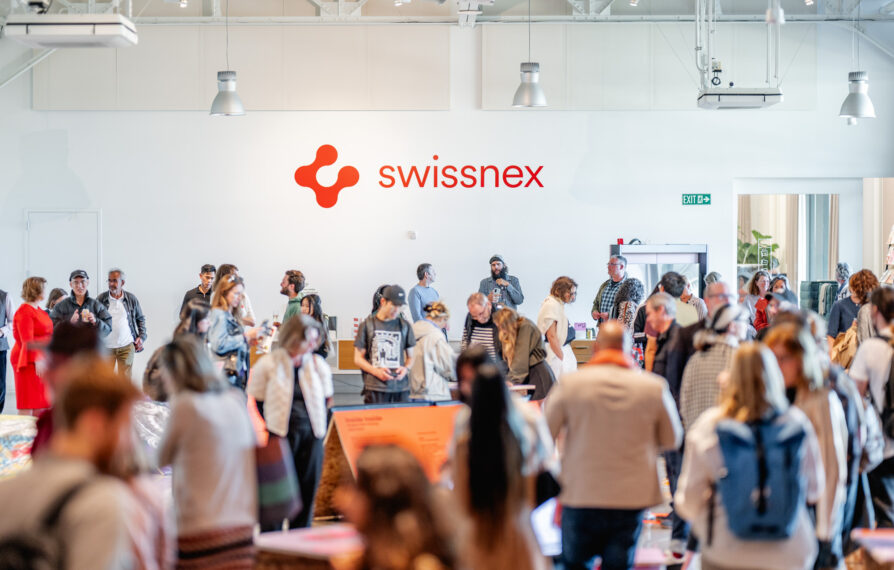 A vibrant gathering at Swissnex featuring a crowd of attendees exploring exhibits and networking in a bright, modern space. The Swissnex logo is prominently displayed on the wall, emphasizing the innovative and collaborative atmosphere. Attendees engage with displays, converse in small groups, and move throughout the open area, which is illuminated by natural light and modern overhead fixtures. The event radiates energy, creativity, and a sense of community, showcasing the spirit of innovation and exchange.