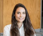 The uploaded image shows a woman with long, dark brown hair, wearing a white, detailed blouse with lace-like patterns and standing confidently with her arms crossed. She has a warm smile, and the background features a modern interior setting with a wooden wall.