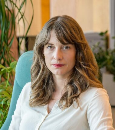 The image features a woman with long, wavy, light brown hair sitting in a teal chair. She is wearing a white blouse and has a confident expression with a slight smile. The background includes lush green indoor plants and softly lit furniture, creating a calm and professional setting.