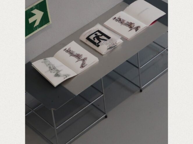 An overhead view of a minimalist display table featuring open sketchbooks and printed materials. The books showcase abstract and artistic designs, including calligraphy-like figures and stylized imagery. The table has a sleek, industrial aesthetic with metal legs and a gray top, positioned against a neutral wall. A green exit sign with an arrow adds a functional and contextual element to the otherwise artistic and curated scene, suggesting an exhibition or creative workspace.
