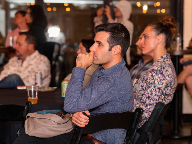 A photo capturing a moment of focused engagement during a Swissnex event on November 6, 2024. The audience members sit attentively, with the foreground highlighting a man in a blue shirt thoughtfully resting his hand against his chin. The dimly lit room and the warm glow of string lights create an intimate and reflective atmosphere. Various individuals in the background add a sense of community and participation, with drinks and casual seating further emphasizing the informal yet professional setting.
