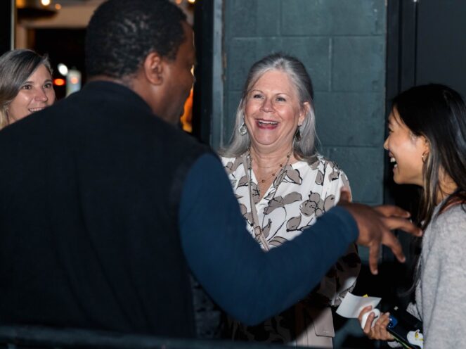 This photo captures a lively and warm interaction during the Swiss Startup Night event on November 6, 2024. A group of attendees is seen engaging in conversation, with expressions of joy and laughter lighting up the scene. The central figure, a woman in a patterned blouse, exudes happiness as she shares in the moment. The setting, slightly dim and casual, complements the relaxed and friendly atmosphere of the networking event. The genuine smiles and animated gestures highlight the collaborative and celebratory spirit of the evening.