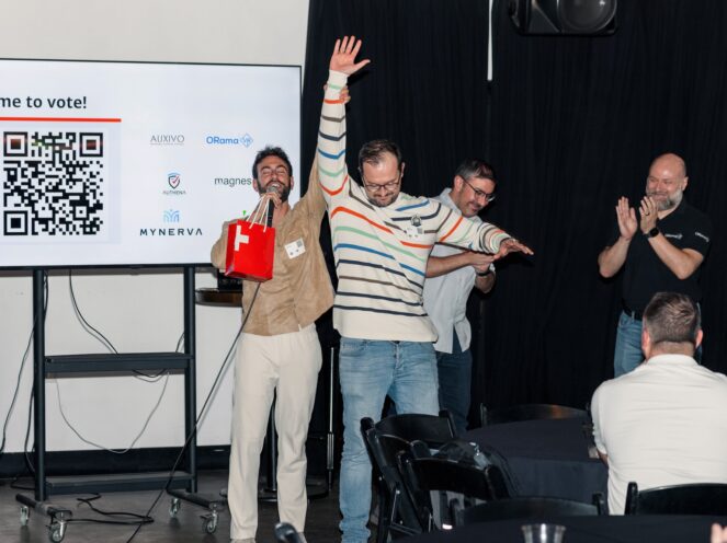 This photo showcases a lively moment from the Swiss Startup Night event on November 6, 2024. Two enthusiastic participants stand center stage, one raising a red Swiss-themed gift bag and microphone, while the other lifts an arm in celebration. They are surrounded by applause and smiles from the audience and fellow participants, reflecting the celebratory atmosphere. The screen in the background displays a QR code and logos of startups, emphasizing the interactive and dynamic nature of the event, likely connected to a voting session for startup presentations or awards.