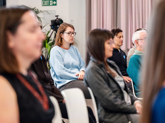 An audience seated in a conference or seminar setting, participating in a guided meditation session. The focus is on a woman in a light blue sweater and glasses, sitting in the center, appearing calm and reflective. Other attendees, both in focus and blurred in the foreground and background, include men and women of various ages, many with eyes closed or serene expressions. A camera on a tripod and an 