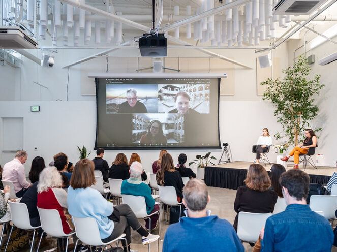 A hybrid panel discussion event featuring a live audience and virtual participants displayed on a large screen. The stage includes two women seated on modern black chairs, engaged in conversation, while the screen behind them shows three remote speakers in a video call, each in different settings. The audience, seated in white chairs, watches attentively in a spacious, well-lit room with high ceilings, modern lighting fixtures, and greenery, creating a contemporary and professional atmosphere. The setup highlights the integration of in-person and virtual collaboration.