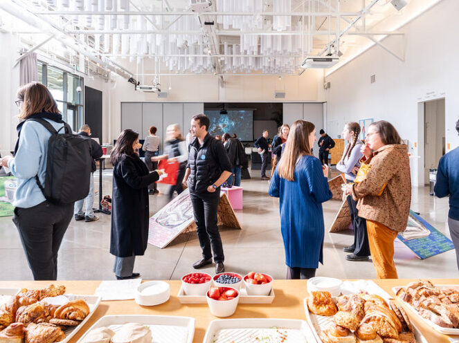 A networking event in a bright, spacious venue featuring attendees engaged in conversations. The foreground showcases a buffet table with a variety of foods, including croissants, bread, fruits, and spreads, adding a welcoming touch. In the background, participants interact near art or informational displays, with a large screen visible against the far wall. The high ceilings, modern lighting fixtures, and large windows create an open and contemporary atmosphere, blending professional and casual elements for a vibrant social gathering.