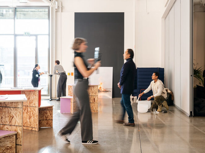 A creative and interactive space within a modern venue, featuring attendees engaging with installations and each other. In the foreground, a person walks past holding a camera or device, while others are seen conversing and exploring. One man is seated on the floor near a sculptural or artistic piece, creating a contemplative moment. The room includes modular wooden tables and bright lighting from large windows, with a minimalist and industrial design that emphasizes openness and collaboration. The setting conveys a mix of art, innovation, and social interaction.