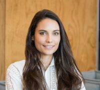 The uploaded image shows a woman with long, dark brown hair, wearing a white, detailed blouse with lace-like patterns and standing confidently with her arms crossed. She has a warm smile, and the background features a modern interior setting with a wooden wall.