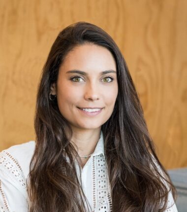 The uploaded image shows a woman with long, dark brown hair, wearing a white, detailed blouse with lace-like patterns and standing confidently with her arms crossed. She has a warm smile, and the background features a modern interior setting with a wooden wall.