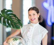 A vibrant and creative portrait of Gwenaëlle Gisiger, exuding confidence and style. She is dressed in a crisp white blouse with ruffle details, holding a reflective disco ball that catches light and adds a playful touch. A large green monstera leaf adds a natural element to the composition, while a neon light in the softly blurred background enhances the modern and dynamic feel of the image. The lighting highlights her cheerful smile and polished appearance, creating an inviting and professional aesthetic.