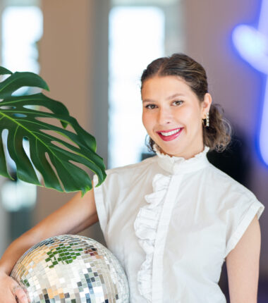 A vibrant and creative portrait of Gwenaëlle Gisiger, exuding confidence and style. She is dressed in a crisp white blouse with ruffle details, holding a reflective disco ball that catches light and adds a playful touch. A large green monstera leaf adds a natural element to the composition, while a neon light in the softly blurred background enhances the modern and dynamic feel of the image. The lighting highlights her cheerful smile and polished appearance, creating an inviting and professional aesthetic.
