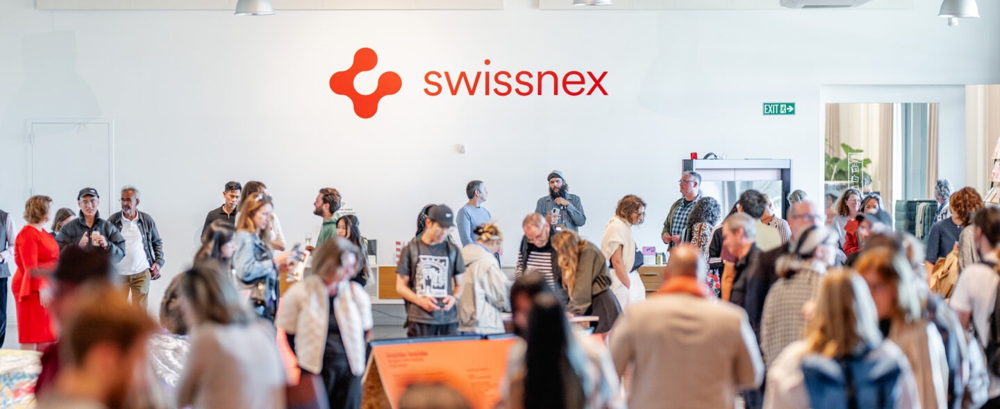 A vibrant gathering at Swissnex featuring a crowd of attendees exploring exhibits and networking in a bright, modern space. The Swissnex logo is prominently displayed on the wall, emphasizing the innovative and collaborative atmosphere. Attendees engage with displays, converse in small groups, and move throughout the open area, which is illuminated by natural light and modern overhead fixtures. The event radiates energy, creativity, and a sense of community, showcasing the spirit of innovation and exchange.
