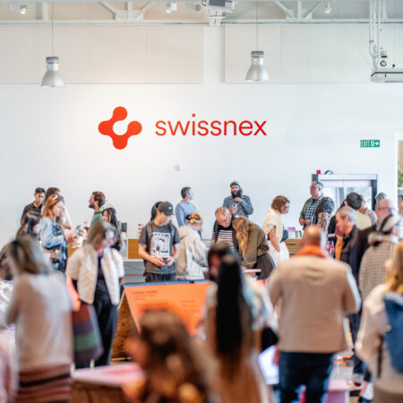 A vibrant gathering at Swissnex featuring a crowd of attendees exploring exhibits and networking in a bright, modern space. The Swissnex logo is prominently displayed on the wall, emphasizing the innovative and collaborative atmosphere. Attendees engage with displays, converse in small groups, and move throughout the open area, which is illuminated by natural light and modern overhead fixtures. The event radiates energy, creativity, and a sense of community, showcasing the spirit of innovation and exchange.