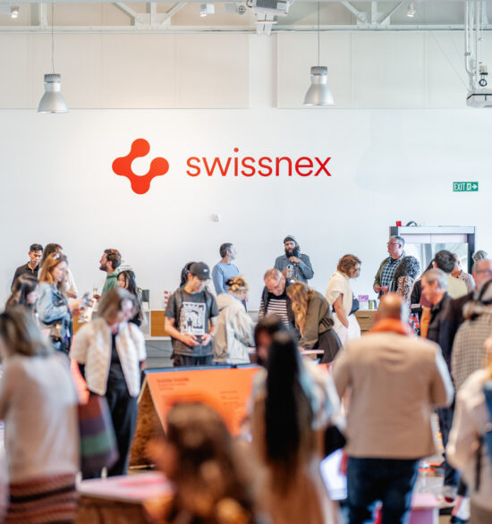 A vibrant gathering at Swissnex featuring a crowd of attendees exploring exhibits and networking in a bright, modern space. The Swissnex logo is prominently displayed on the wall, emphasizing the innovative and collaborative atmosphere. Attendees engage with displays, converse in small groups, and move throughout the open area, which is illuminated by natural light and modern overhead fixtures. The event radiates energy, creativity, and a sense of community, showcasing the spirit of innovation and exchange.