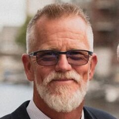 This is a portrait of a man with a white beard, short cropped hair, and glasses. He is dressed in formal attire, wearing a suit and tie. The blurred background suggests an outdoor setting, perhaps near a waterfront or urban area. The overall image conveys professionalism with a casual touch due to the natural lighting and setting.