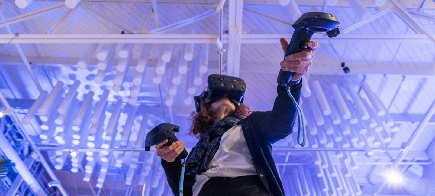 An immersive moment showcasing a person wearing a VR headset and holding motion controllers, engaged in a virtual reality experience. The low-angle shot highlights their active participation, with their arms extended in a dynamic pose. The setting features a modern ceiling with artistic cylindrical light fixtures, illuminated in soft blue hues, creating a futuristic and engaging atmosphere. The combination of technology and creativity emphasizes innovation and exploration.