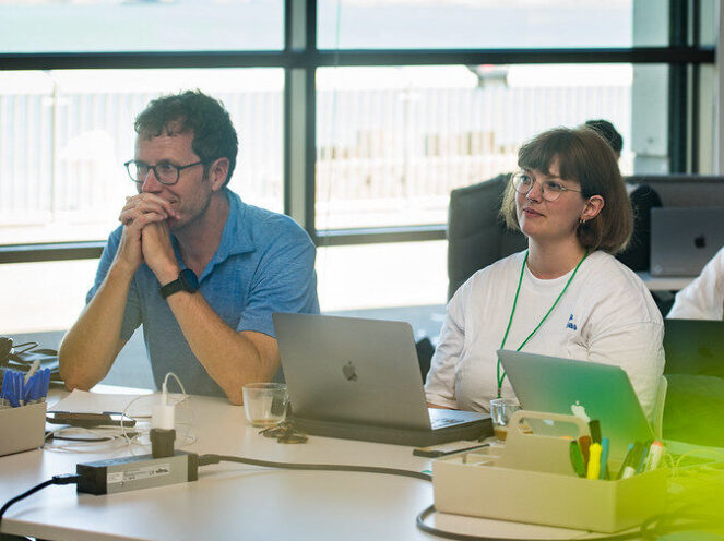 A focused moment during a workshop featuring two participants seated at a table with laptops and office supplies. The man on the left, wearing a blue shirt and glasses, leans forward with hands clasped in thought, while the woman on the right, wearing a white shirt and glasses, listens attentively. The bright space features large windows with a view of a waterfront, adding natural light and a scenic backdrop. The atmosphere suggests a collaborative and educational setting, fostering engagement and creativity.