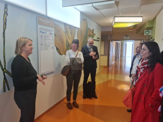 A group of people standing in a hospital hallway with a guide explaining something to them. The walls are decorated with floral murals, and a sign indicates the location as UCSF Medical Center. The floor is brightly colored, creating a welcoming atmosphere.