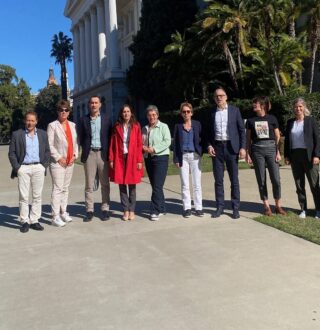 A group of ten people posing for a photo outdoors on a sunny day in front of a white neoclassical building surrounded by palm trees and lush greenery. The individuals are standing on a paved walkway, dressed in a mix of formal and casual attire, and the background shows a clear blue sky and distant buildings.